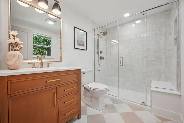 Upper Arlington primary bath with an oak vanity, a brass mirror, a glass walk-in shower and a checkerboard floor